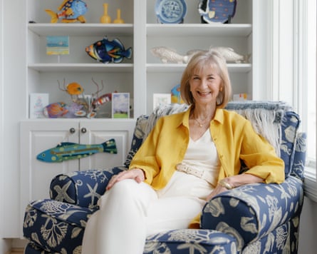 Author Lindsay Gibson in a yellow shirt sits on a blue patterned armchair with shelves of decorative fish behind her.
