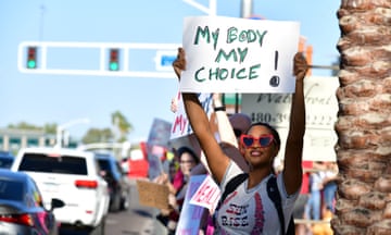 A woman in heart-shaped glasses holds a "My Body My Choice" sign at a traffic intersection.