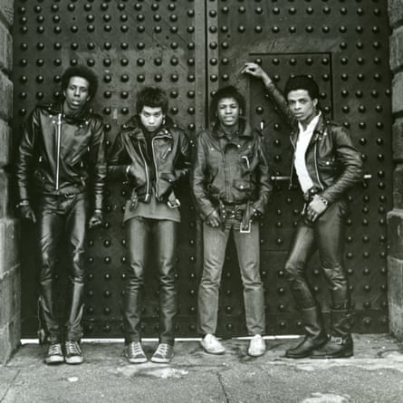 A black-and-white photo of four young, skinny Black men in leather jackets and jeans or leather pants, standing leaning against a tall studded double door, as if for a roadway behind them.