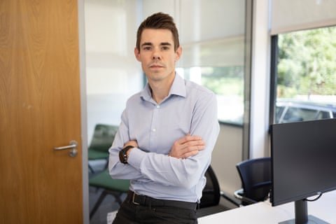 A man wearing a shirt with his arms folded standing next to a desk with a computer on it.