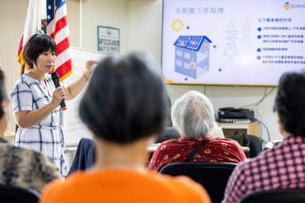 A woman speaks into a mic before an audience of elderly people.