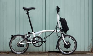 An electric Brompton standing against pale blue wooden cladding
