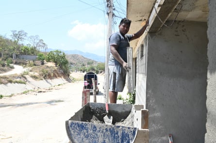 A man holds what appears to be a piece of wood, the other hand is on the lintel of a concrete building. A trough of what appears to be cement and a trowel sit next to him