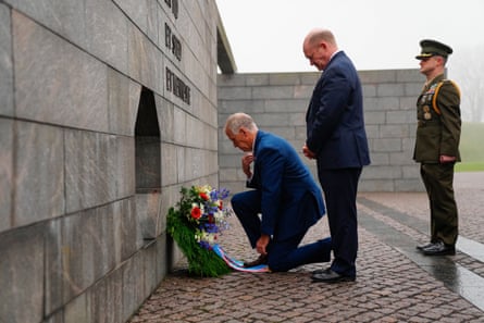 Two men in blue suits flanked by a man in a green military uniform standing at attention behind them. One of the men in a blue suit kneels at a cement wall before a wreath.