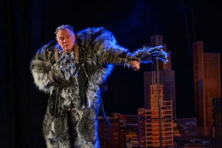 Stewart Lee performing on stage, wearing a full-body, grey furry werewolf outfit and leaning the left clawed hand of the outfit on a microphone on a stand