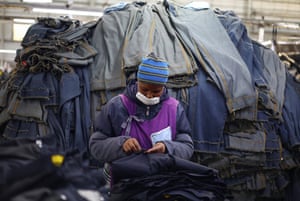 A worker, wearing a wooly hat and face mask, repairs clothing in front of a large mound of clothes