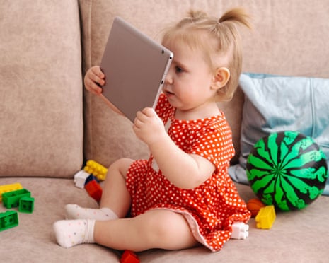 A toddler in a red polka dot dress holds a tablet while sitting on a beige sofa surrounded by toys