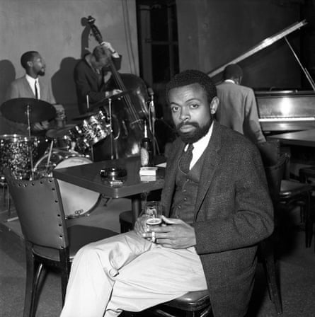 A man sits holding a drink as a jazz band plays behind him