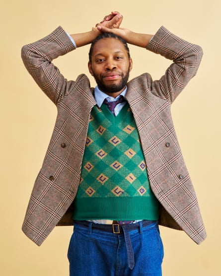 Jeremy O Harris standing with hands resting on his head. He is wearing jeans, a tweed blazer, and a knitted vest over a shirt and tie