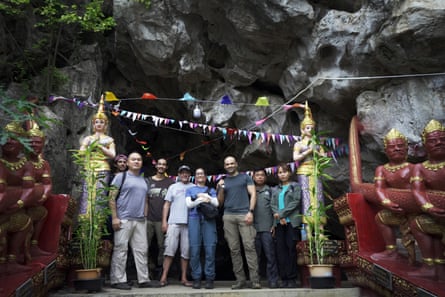 A group of people stand at the mouth of a cave between two Buddha statues strung with flags.