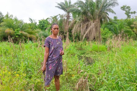 An African woman stands in long grass holding a machete and wearing a floral purple dress, with trees in the distance and greenery all around.
