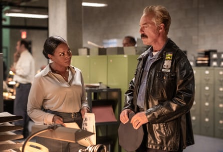 A policeman in a black leather jacket talks to woman standing over a desk.