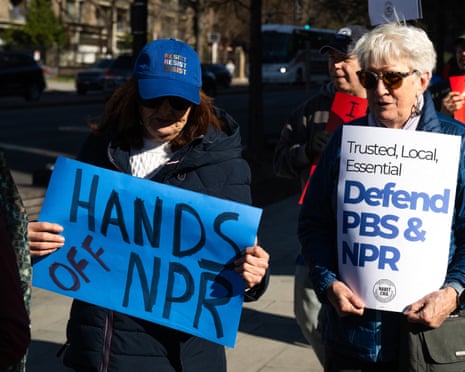 Two people holding signs reading ‘hands off NPR’ and ‘defend PBS & NPR’ at a protest