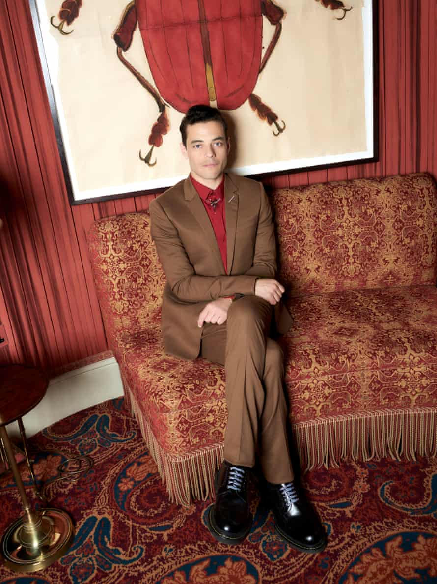 Rami Malek in a brown suit, red shirt and black shoes, sitting cross-legged on red patterned upholstery