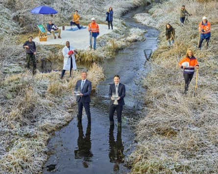 Two men in suits standing a stream holding glass bowls as other people, some in hi-vis jackets and one in a white lab coat stand on the grassy banks