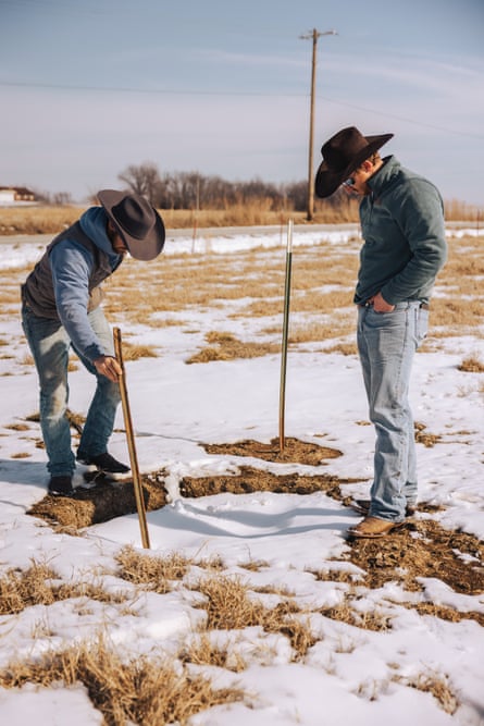 two men in cowboy hats, blue jeans, and boots examine a small sink hole filled with snow