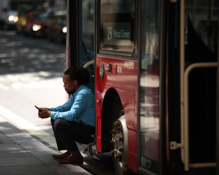 A bus driver sits in the doorway of his bus, looking at his smartphone.