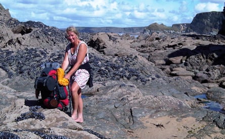 Raynor Winn in sundress on a rocky coastline, with big rucksack