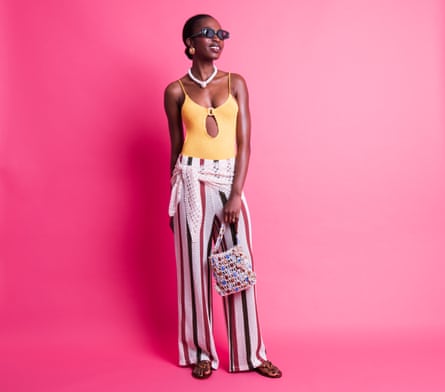 A woman in a yellow one-piece swimsuit, cream crotchet stripe trousers, crotchet scarf round her waist, a cream beaded flower rope bag, sandals and sunglasses