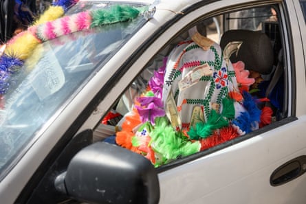 Someone with their head covered in a cloth with money pinned to it and garlands of plastic flowers around their neck sits in the front passenger seat of a car.