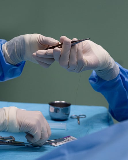 The left hand of the doctor passes a surgical instrument to the right hand of his assistant; both are wearing white gloves and blue scrubs. The doctor’s other hand is lowered towards another instrument on a blue surface.