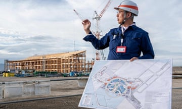 JA Japanese man in a hard hat holds a large map and gestures to a construction site in the background with a big wooden structure and a giant crane