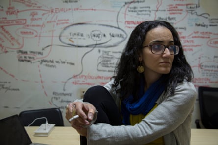 Lina Attalah holds a meeting in the Mada Masr office in 2015. She has long dark hair and wears glasses, and is holding a cigarette as she sits on a desk in front of a whiteboard scribbled with notes.