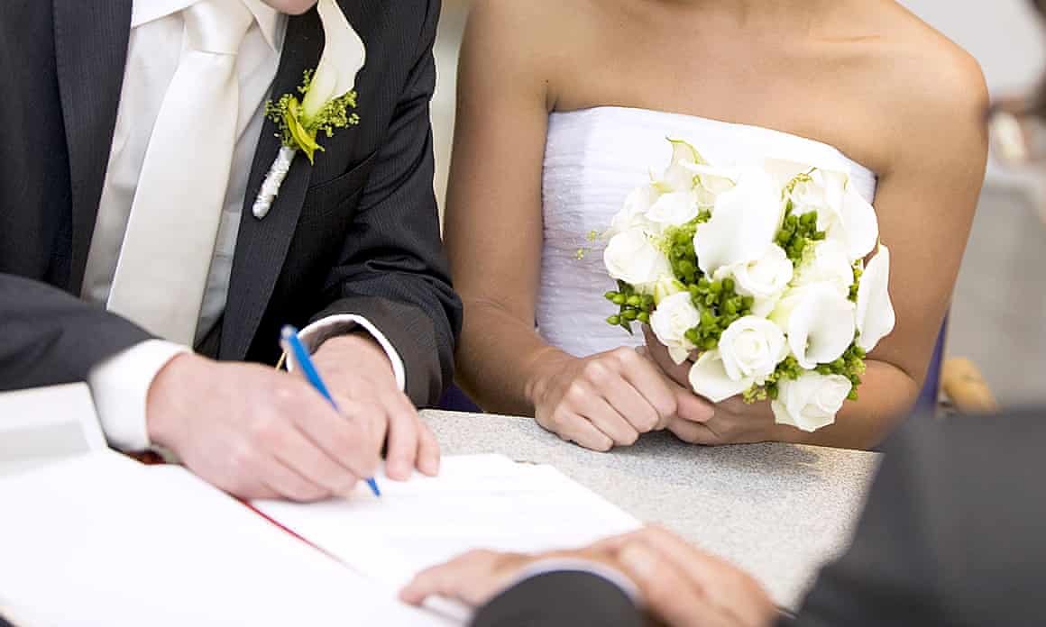 A couple sit together in their wedding outfits as the man signs a piece of paper