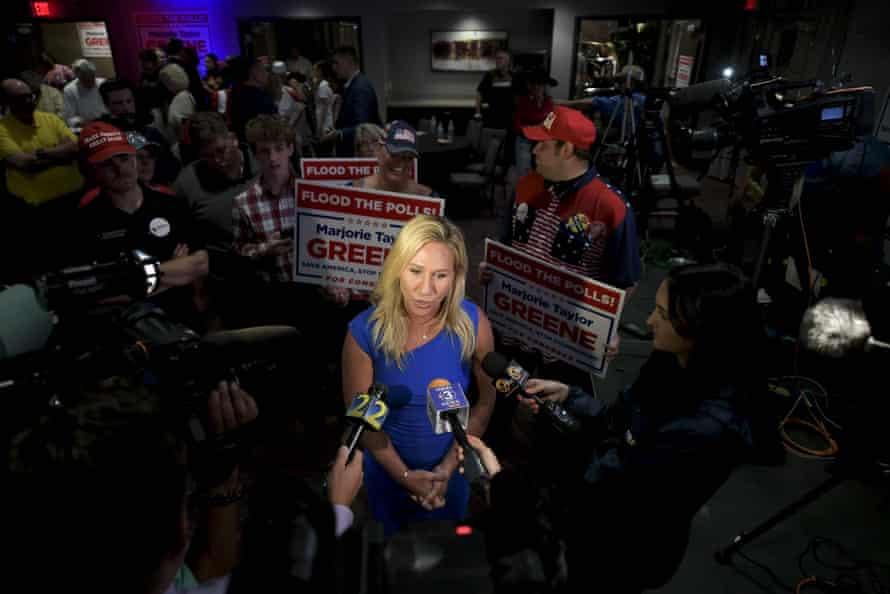Marjorie Taylor Greene is seen from above, standing in a crowd of supporters while talking into two microphones.