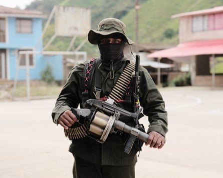 A man in military uniform and balaclava walks across a sports court holding a grenade launcher