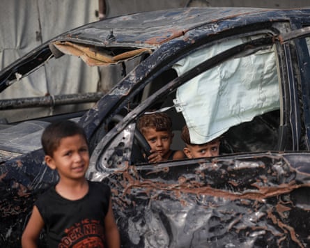 Two young boys hide in a burnt-out car as another stands in front of it