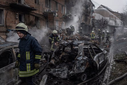 Firefighters walk past the burnt-out husk of a car at the side of a road