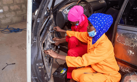 Two young women in brightly coloured overalls and headscarves work on a car door