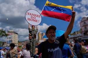 A man holds a doll of Hugo Chavez and a siign that reads “Yankee go home!”. Behind him, someone waves a Venezuela flag