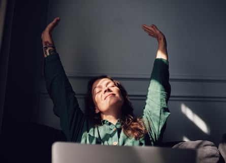 A young woman stretches her arms right up at a desk, in front of a laptop