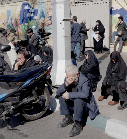 An elderly man sits on the kerb with his head in his hands, looking sad