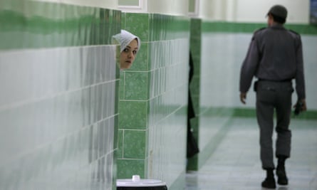 A woman with a white head covering peers from behind a wall as a guard walks by