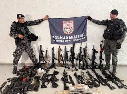 Two men in military clothing stand over a haul of weapons and hold a flag that says Policia Militar