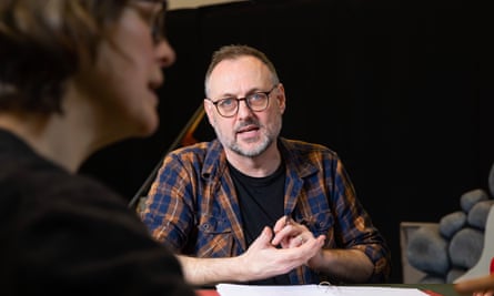 Simon Harvey, a middle aged man with a small beard and brown hair, wearing glasses, an open shirt and T-shirt, talking to people at a table