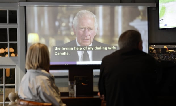 Žena a muž při pohledu zezadu sledují krále Charlese, jak mluví na obrazovce v hospodě v centru Londýna