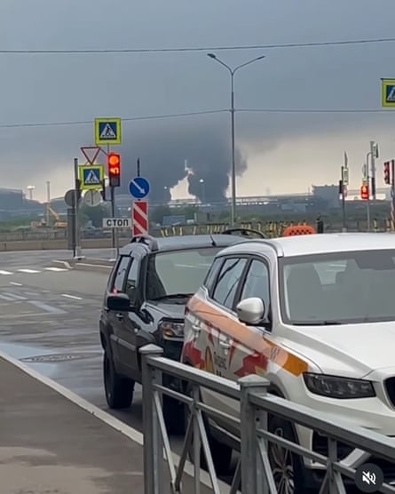 Two plumes of black smoke visible on the horizon behind cars parked in a Russian industrial port zone.