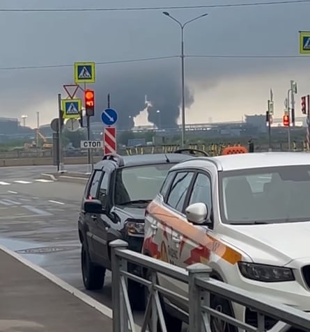 Two plumes of black smoke visible on the horizon behind cars parked in a Russian industrial port zone.