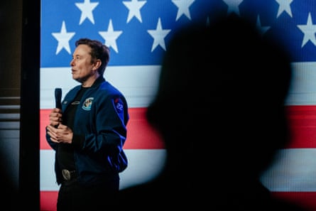 A man on stage speaks into a microphone with the US flag behind him.