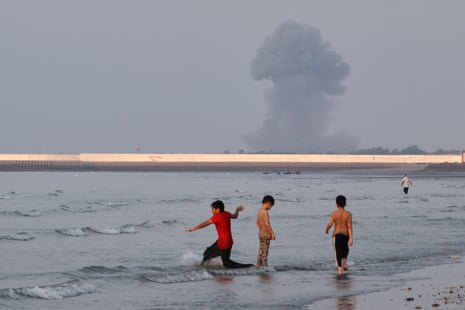 Three boys play in the shallow waters of the strait of Hormuz, as a plume of smoke rises from an explosion in the background off the Iranian city of Bandar Abbas.