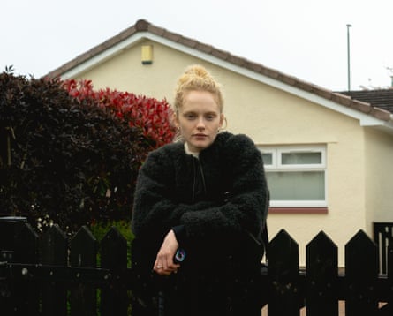 A young woman stands leaning on a front gate
