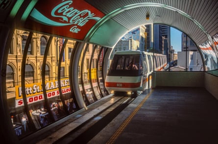 A monorail station with a train arriving to the platform, with a CocaCola sign visible and an overall 80s feel to the picture.