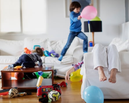 Playful chaos as children jump from sofa to sofa, surrounded by toys and balloons in a messy living room. Meanwhile, their tired mother reclines on the couch