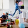 Playful chaos as children jump from sofa to sofa, surrounded by toys and balloons in a messy living room. Meanwhile, their tired mother reclines on the couch