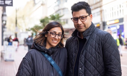 Close up of couple smiling in black coats and spectacles standing in a high street.