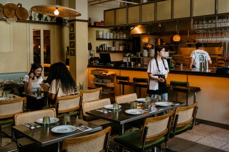 Staff and diners in a restaurant with wooden chairs, set tables, and an open kitchen area